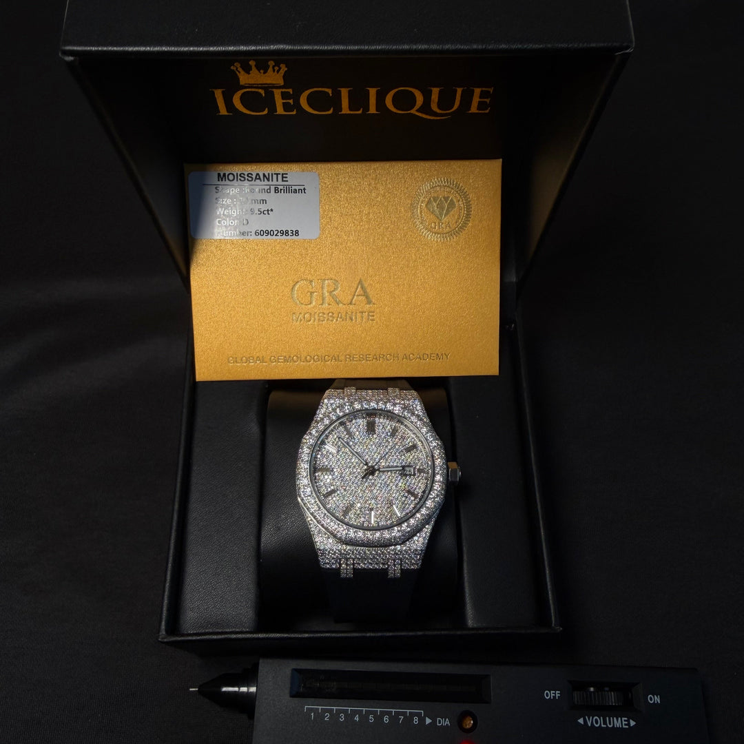 A luxury watch presented in an open black Iceclique box with a gold certificate insert, displayed above a black diamond selector tool on a black background. The watch has a diamond-encrusted bezel, a silver-toned dial, and a dark strap.